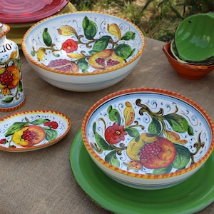 K&ouml;nnte beinhalten: Eine Sammlung handbemalter Keramikgeschirr mit Granatapfel-Design. Das Set umfasst Schalen, Teller und eine &Ouml;lflasche. Die Farbpalette umfasst Wei&szlig;, Blau, Gelb, Rot und Gr&uuml;n. Auf der &Ouml;lflasche steht das Wort "Olio".