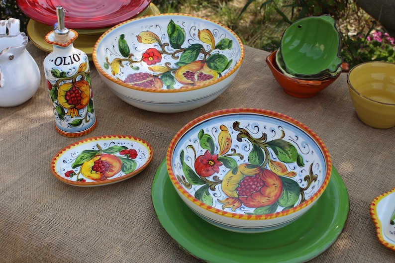 K&ouml;nnte beinhalten: Eine Sammlung handbemalter Keramikgeschirr mit Granatapfel-Design. Das Set umfasst Schalen, Teller und eine &Ouml;lflasche. Die Farbpalette umfasst Wei&szlig;, Blau, Gelb, Rot und Gr&uuml;n. Auf der &Ouml;lflasche steht das Wort "Olio".