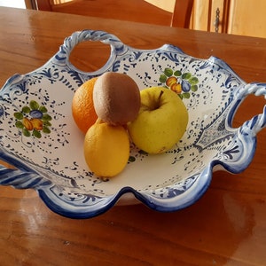 K&ouml;nnte beinhalten: Eine wei&szlig;e Keramikservierschale mit blau-wei&szlig;em Blumendesign, die eine Orange, Kiwi, Zitrone und einen gelben Apfel enth&auml;lt. Die Schale hat Griffe und einen gewellten Rand. Das Obst ist frisch und farbenfroh.