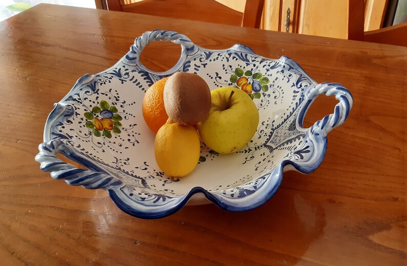 K&ouml;nnte beinhalten: Eine wei&szlig;e Keramikservierschale mit blau-wei&szlig;em Blumendesign, die eine Orange, Kiwi, Zitrone und einen gelben Apfel enth&auml;lt. Die Schale hat Griffe und einen gewellten Rand. Das Obst ist frisch und farbenfroh.