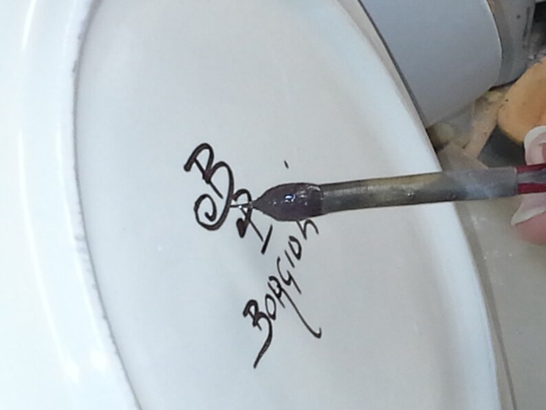 K&ouml;nnte beinhalten: Nahaufnahme einer wei&szlig;en Keramikoberfl&auml;che mit schwarzer Schreibschrift und einem Metallstift mit violetter Tinte. Der Stift befindet sich &uuml;ber der Oberfl&auml;che, im Begriff zu schreiben. Der Text lautet "B. I. BORGION".