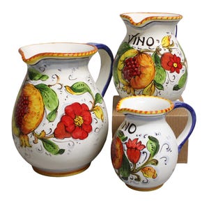 Puede incluir: Tres jarras de cerámica blanca con coloridos diseños florales y frutales. Las jarras tienen asas azules y bordes con detalles amarillos y naranjas. La palabra "VINO" está escrita en dos de las jarras.