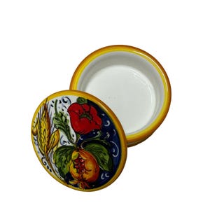 Può includere: Un portagioie rotondo in ceramica con coperchio rimovibile. Il coperchio presenta un disegno dipinto a mano di un papavero rosso, foglie verdi e spighe di grano gialle su sfondo blu scuro. Il bordo del coperchio e la base sono rifiniti in giallo.