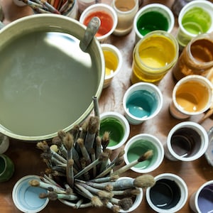 K&ouml;nnte beinhalten: Draufsicht auf einen K&uuml;nstlerarbeitsplatz mit einer gro&szlig;en Wassersch&uuml;ssel, Pinseln und verschiedenen Beh&auml;ltern mit bunten Farben. Die Farben umfassen Gr&uuml;nt&ouml;ne, Gelb, Orange und Blau, was auf einen Schwerpunkt auf Malerei und Kunstbedarf hindeutet.