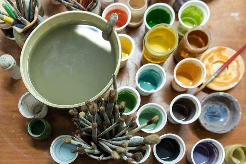 K&ouml;nnte beinhalten: Draufsicht auf einen K&uuml;nstlerarbeitsplatz mit einer gro&szlig;en Wassersch&uuml;ssel, Pinseln und verschiedenen Beh&auml;ltern mit bunten Farben. Die Farben umfassen Gr&uuml;nt&ouml;ne, Gelb, Orange und Blau, was auf einen Schwerpunkt auf Malerei und Kunstbedarf hindeutet.