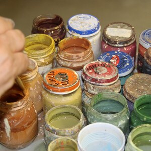 May include: A selection of open glass jars filled with different colours of paint. The jars have metal lids, some with labels. The paint colours include yellow, brown, green, and blue. A hand is visible, stirring the paint in one of the jars.