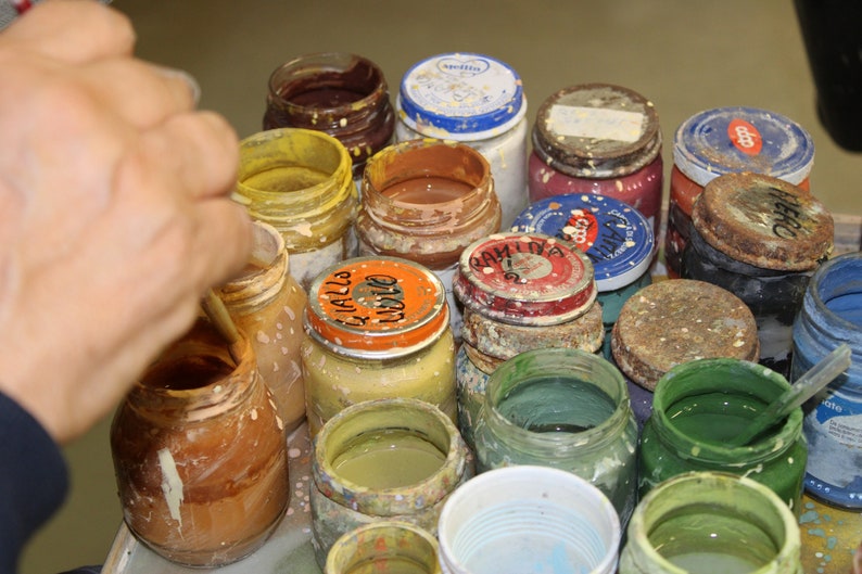 May include: A selection of open glass jars filled with different colours of paint. The jars have metal lids, some with labels. The paint colours include yellow, brown, green, and blue. A hand is visible, stirring the paint in one of the jars.