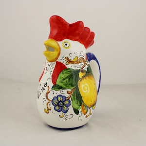 Peut inclure: Une cruche en céramique blanche en forme de coq avec une crête et une barbe rouges. La cruche est décorée d'un citronnier, de fleurs et du mot "Vino" en cursive.