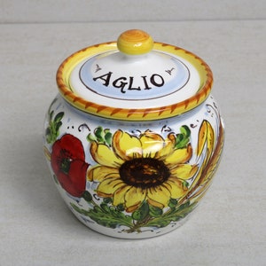 May include: A ceramic jar with a lid, featuring a hand-painted design. The jar is adorned with a large yellow sunflower, a red poppy, and green leaves. The lid has the word "AGLIO" and a yellow knob. The rim is yellow and brown.