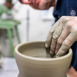 K&ouml;nnte beinhalten: Nahaufnahme der H&auml;nde eines T&ouml;pfers, die mit nassem Ton bedeckt sind und eine hellbeige Keramikvase auf einer T&ouml;pferscheibe formen. Der T&ouml;pfer tr&auml;gt ein dunkelblaues Hemd. Im Hintergrund befindet sich eine gr&uuml;ne T&ouml;pferscheibe.