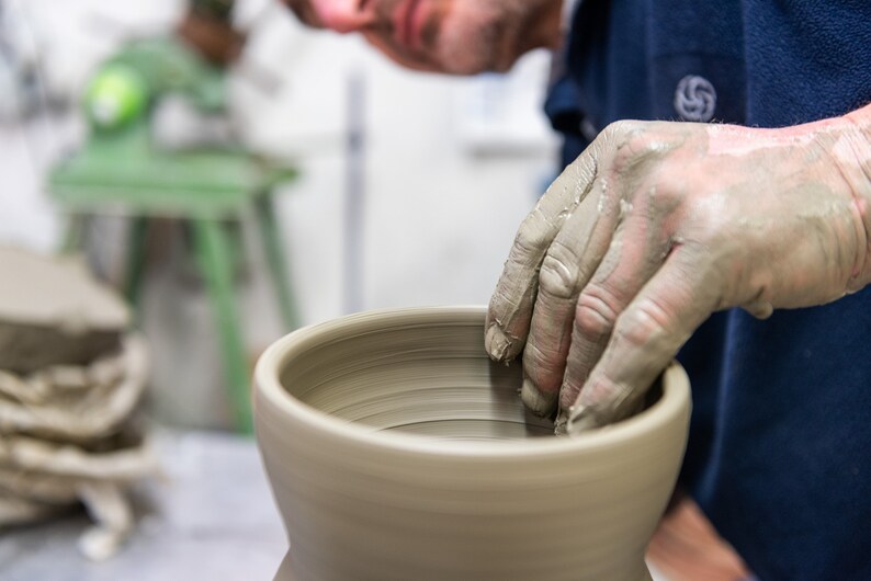 K&ouml;nnte beinhalten: Nahaufnahme der H&auml;nde eines T&ouml;pfers, die mit nassem Ton bedeckt sind und eine hellbeige Keramikvase auf einer T&ouml;pferscheibe formen. Der T&ouml;pfer tr&auml;gt ein dunkelblaues Hemd. Im Hintergrund befindet sich eine gr&uuml;ne T&ouml;pferscheibe.