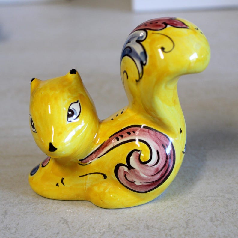 May include: A ceramic squirrel figurine in a bright yellow hue. The squirrel has black eyes and ears, with decorative swirls in burgundy, blue, and black. The figurine is a decorative item.