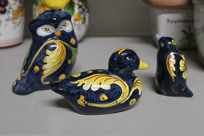 May include: Three ceramic figurines: an owl, a duck, and a bird. The owl and bird are upright, while the duck is in a resting position. All are navy blue with yellow and white floral designs and yellow beaks.