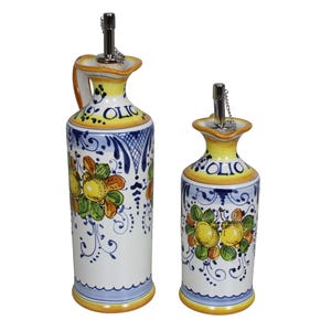 Könnte beinhalten: Zwei Olivenölspender aus Keramik. Die Spender sind weiß mit gelben und blauen Akzenten. Sie weisen ein Blumendesign mit gelben und grünen Früchten auf. Das Wort "OLIO" steht auf der Oberseite jedes Spenders.