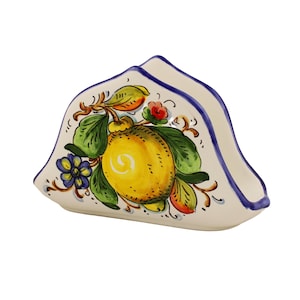 May include: A ceramic napkin holder with a hand-painted lemon design. The holder is off-white with a blue trim and features a large yellow lemon, green leaves, and small red and blue flowers. The design is in a classic Italian style.