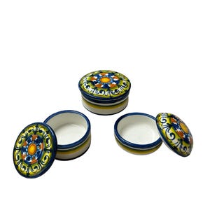 Può includere: Tre scatole portagioie rotonde in ceramica con coperchio, caratterizzate da un motivo dipinto a mano in blu, giallo, verde e arancione. I coperchi presentano un motivo floreale centrale circondato da volute, con strisce orizzontali.