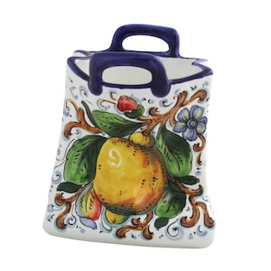 Puede incluir: Un portautensilios de cerámica blanca con asa y borde azules. El portautensilios está decorado con una fruta amarilla, hojas verdes y flores azules y rojas. El diseño está pintado a mano con detalles marrones.