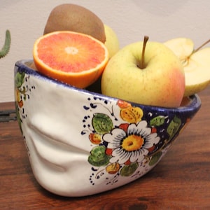 Può includere: Una ciotola in ceramica bianca a forma di volto umano, decorata con motivi floreali dipinti a mano in blu, giallo, verde e arancione. La ciotola è riempita di frutta fresca, tra cui un'arancia rossa tagliata a metà, una mela e un kiwi.