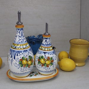 Puede incluir: Dos botellas de aceite de oliva de cerámica con diseños florales azules y blancos y detalles dorados. Las botellas están etiquetadas como "Olio" y "Aceto". Las botellas están sobre una bandeja ovalada blanca con limones delante.
