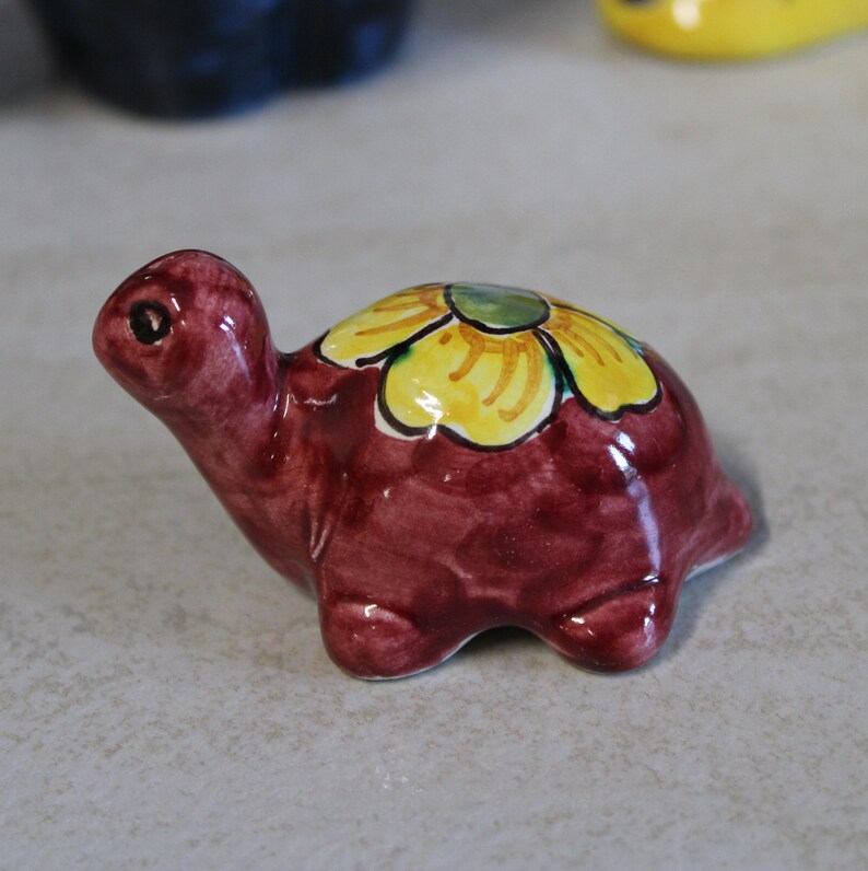 May include: A ceramic turtle figurine in a glossy burgundy finish. The turtle's shell is decorated with a yellow and green flower design. The figurine is a decorative item.