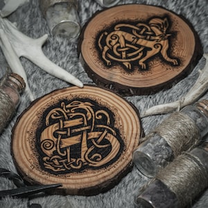 May include: Two wooden coasters with intricate black and white designs of Celtic knots and animals. The coasters are on a gray furry surface with small glass bottles wrapped in twine.