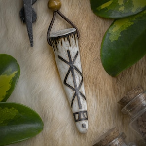 Op de afbeelding: Een cr&egrave;mekleurige hangerketting met een bruin leren koord. De hanger is versierd met een zwart geometrisch ontwerp. De ketting wordt getoond op een getextureerd, lichtgekleurd oppervlak, met groene bladeren en kleine glazen potjes op de achtergrond.