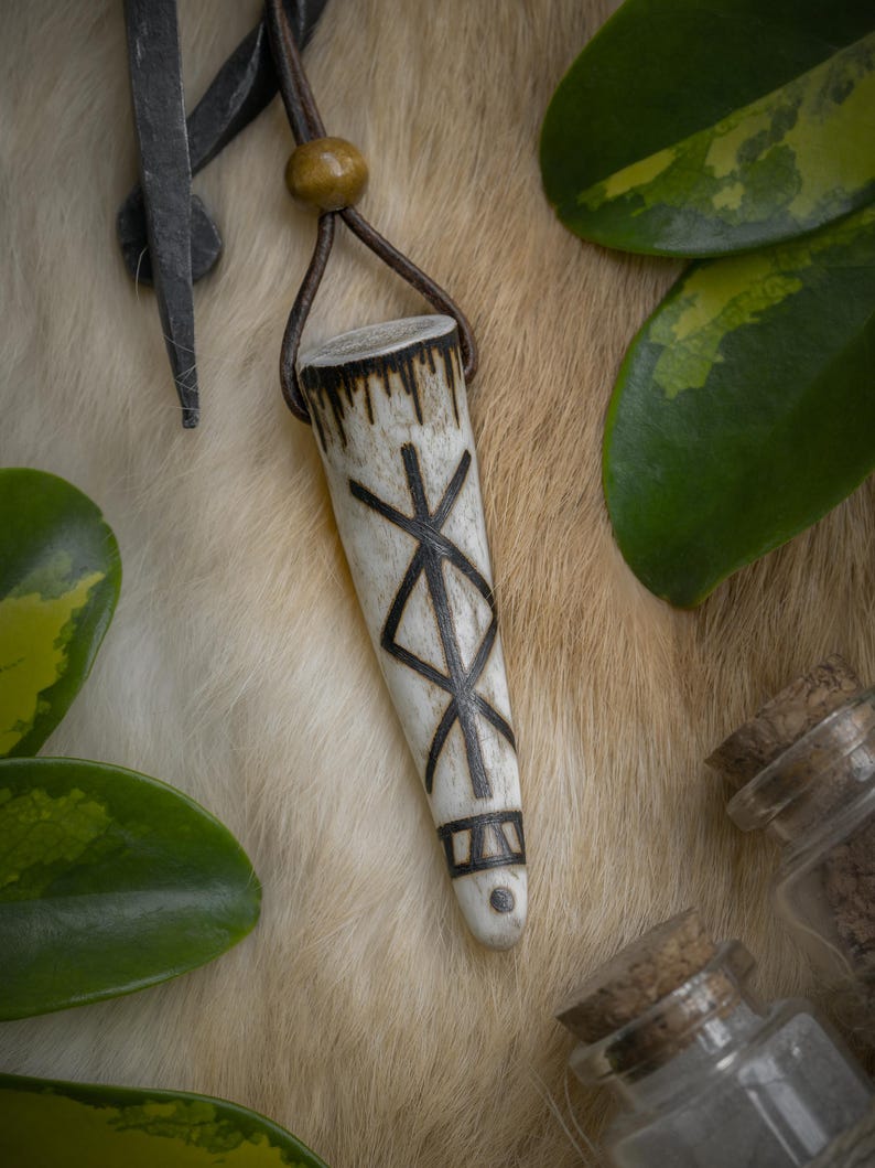 Op de afbeelding: Een cr&egrave;mekleurige hangerketting met een bruin leren koord. De hanger is versierd met een zwart geometrisch ontwerp. De ketting wordt getoond op een getextureerd, lichtgekleurd oppervlak, met groene bladeren en kleine glazen potjes op de achtergrond.