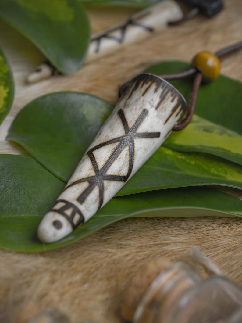 Op de afbeelding: Een cr&egrave;mekleurige hanger met een donkerbruin geometrisch ontwerp en een leren koord. De hanger is kegelvormig en heeft een houten kraal. De achtergrond omvat groene bladeren en een textuur.