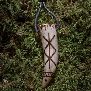 Op de afbeelding: Een hangerketting met een gesneden, gebroken witte hoornhanger met donkerbruine ge&euml;tste symbolen. De hanger hangt aan een donkergrijze leren koord met een houten kraal. De achtergrond is groen mos.