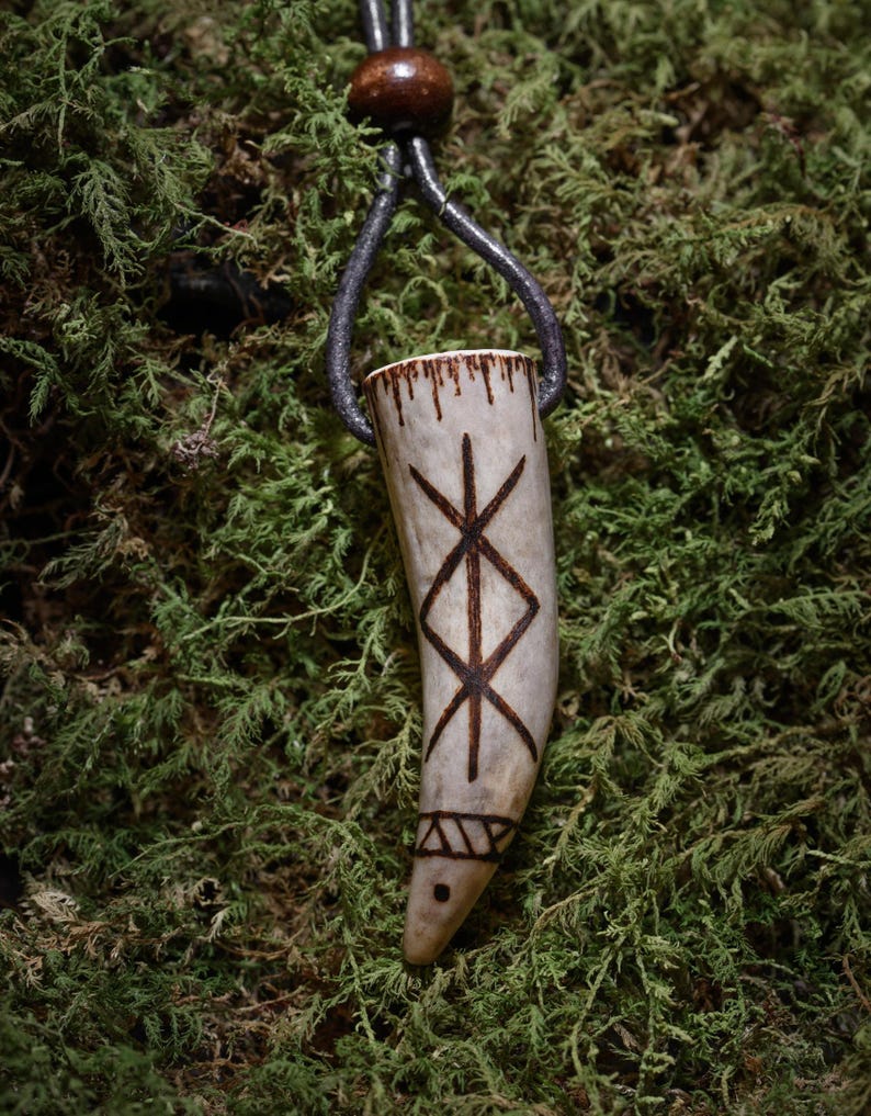 Op de afbeelding: Een hangerketting met een gesneden, gebroken witte hoornhanger met donkerbruine ge&euml;tste symbolen. De hanger hangt aan een donkergrijze leren koord met een houten kraal. De achtergrond is groen mos.