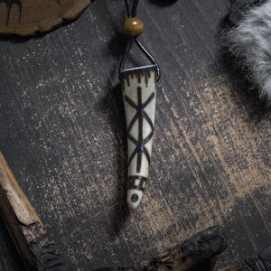 Op de afbeelding: Een cr&egrave;mekleurige hangerketting met een zwart leren koord. De hanger heeft een taps toelopende vorm met een donker, geometrisch ontwerp. De ketting wordt gepresenteerd op een donkere, getextureerde ondergrond met houten accenten.