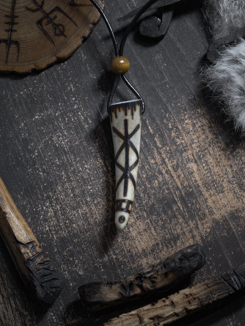 Op de afbeelding: Een cr&egrave;mekleurige hangerketting met een zwart leren koord. De hanger heeft een taps toelopende vorm met een donker, geometrisch ontwerp. De ketting wordt gepresenteerd op een donkere, getextureerde ondergrond met houten accenten.