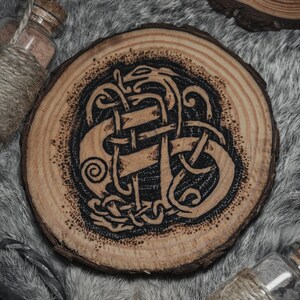 May include: A wooden coaster with a black and brown intricate design of a knotwork pattern. The coaster is round and has a natural wood grain.