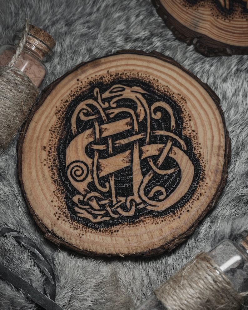 May include: A wooden coaster with a black and brown intricate design of a knotwork pattern. The coaster is round and has a natural wood grain.