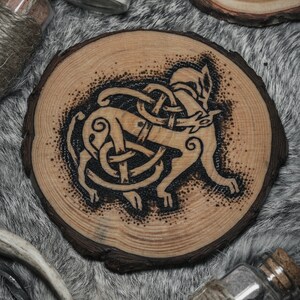 May include: A wooden coaster with a black and white burned design of a wolf in a Celtic knot. The coaster is round and has a natural wood finish.