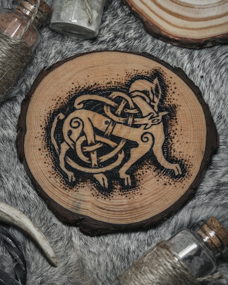 May include: A wooden coaster with a black and white burned design of a wolf in a Celtic knot. The coaster is round and has a natural wood finish.