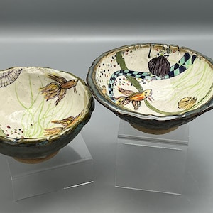 May include: Two ceramic bowls with hand-painted aquatic designs. One bowl features a fish and plant life, while the other has a snake-like creature and fish. Both bowls have a dark rim and sit on clear stands.