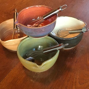 May include: Four ceramic mixing bowls in shades of beige, purple, green, and black, each with a stainless steel whisk. The bowls have a pouring spout. The whisks have a red handle.