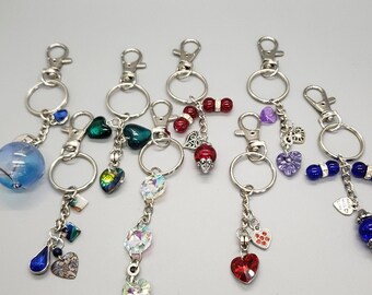 May include: Seven silver keychains with different charms. The charms include a blue glass sphere, green glass hearts, crystal beads, red glass hearts, purple glass hearts, and blue glass beads.