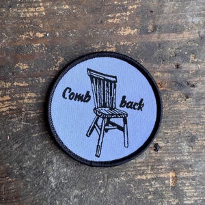May include: A circular embroidered patch with a light blue background and a black stitched border. The patch features a black line art illustration of a wooden chair and the black embroidered text "Comb back" positioned around the chair.