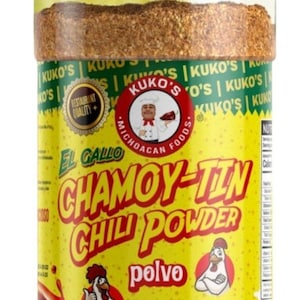 Op de afbeelding: Een gele en rode plastic pot met El Gallo Chamoy-Tin Chilipoeder, met een groene deksel. De pot heeft een cartoonhaan op de voorkant, en de tekst "polvo" is onder de haan gedrukt. De pot is gevuld met een bruin poeder. De pot is 5 oz.
