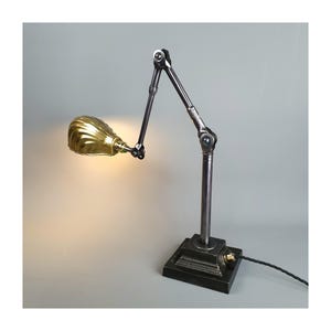 May include: An industrial desk lamp featuring a brass shade and an adjustable arm. The lamp has a dark metal base and arm, with a cord extending from the base. The shade is a warm gold colour.