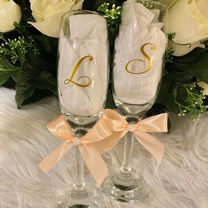 May include: Two champagne flutes with peach-colored bows and gold lettering. The flutes are decorated with the letters "L" and "S".