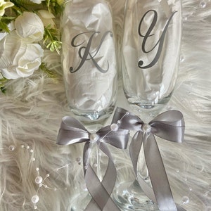 May include: Two champagne flutes with gray satin bows and pearl accents. The flutes are personalized with the letters "K" and "Y" in a cursive font.