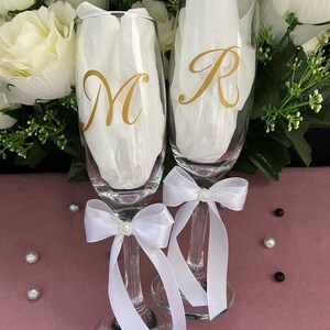 May include: Two champagne flutes with white satin bows and pearl accents. The flutes are personalized with the letters "M" and "R" in gold.