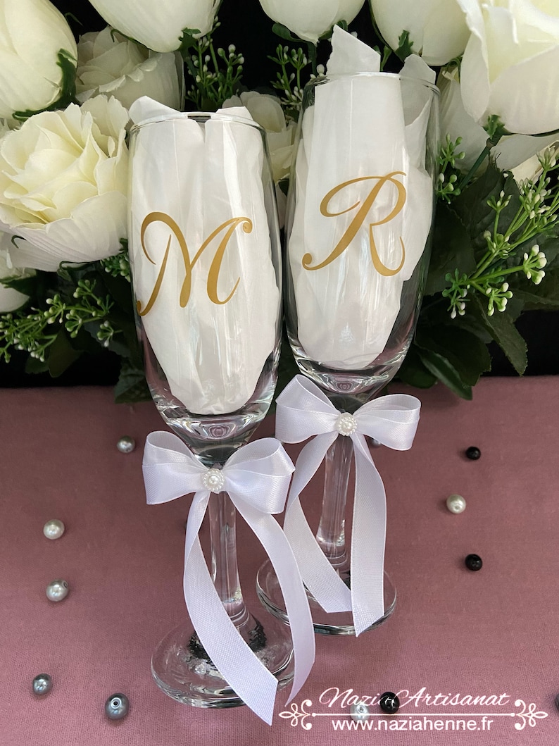 May include: Two champagne flutes with white satin bows and pearl accents. The flutes are personalized with the letters "M" and "R" in gold.