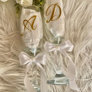 May include: Two champagne flutes with white satin bows and pearl accents. The flutes are decorated with gold vinyl letters, "A" and "D".