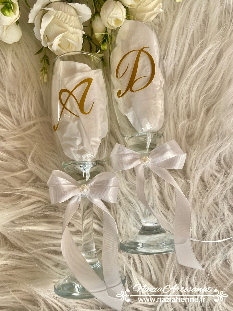 May include: Two champagne flutes with white satin bows and pearl accents. The flutes are decorated with gold vinyl letters, "A" and "D".