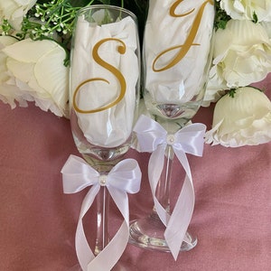 May include: Two champagne flutes with white satin bows and pearl accents. The flutes are decorated with gold vinyl letters, "S" and "J".