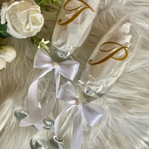 May include: Two champagne flutes with white satin ribbons and pearl accents. The flutes are decorated with gold vinyl letters, one with the letter "J" and the other with the letter "P".